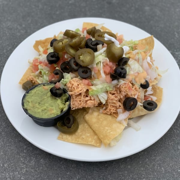 A plate of nachos with shredded chicken, lettuce, tomatoes, black olives, jalapeños, and a side of guacamole.
