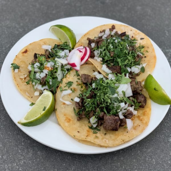 Three street tacos with steak, onions, cilantro, lime wedges, and a radish on a white plate.