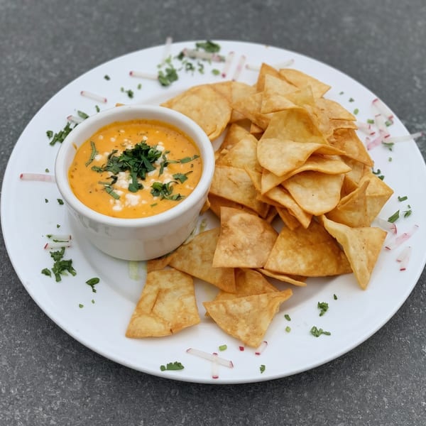 A white bowl of orange queso dip with cilantro and feta, served with tortilla chips.